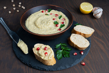 Hummus savory dish made of mashed chickpeas with tahini, lemon juice and garlic decorated with pomegranate seeds on round tray with bread slices and knife on wooden background. Horizontal orientation