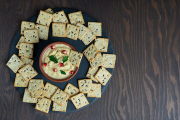 Top view of savory homemade vegetarian hummus sauce made of chickpeas  decorated with pomegranate seeds and parsley served with crispy snack on wooden background at kitchen. Image with copy space
