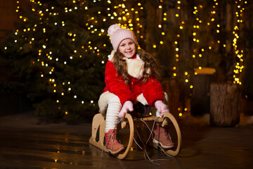 Happy child girl in winter clothes sitting on a wooden sleigh on background and xmas tree on Christmas Eve outdoor.