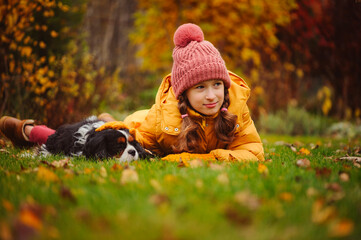 Happy kid girl walking and playing with her cavalier king charles spaniel dog in autumn garden or...