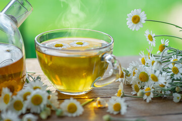 Herbal chamomile tea and chamomile flowers near teapot and tea glass on wooden table. Countryside background.