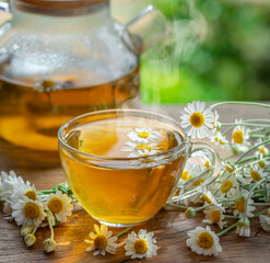 Herbal chamomile tea and chamomile flowers near teapot and tea glass on wooden table. Countryside...