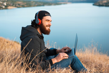 Young bearded man listening to the music and working on the computer on the field near a river
