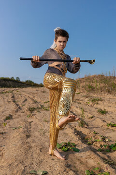 A teenage guy in stylish fantasy clothes in an old middle eastern style richly decorated with gold jewelry and a cane in his hands with a knob on the setting sun on the sand