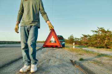 Fototapeta na wymiar Cropped shot of man driver putting red warning triangle or emergency stop sign behind his broken car on the side of the road, his girlfriend waiting in the background