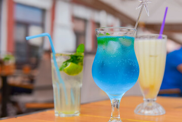 Three fresh juice drinks in tall glasses with plastic straws outside. Milkshake, blue curacao cocktail and mohito.