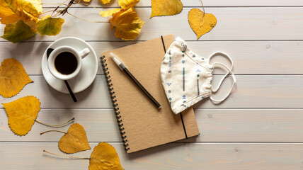 Workspace with notebook, mask, pen, autumn leaves, and coffee cup.