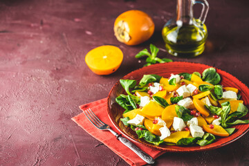Healthy salad with persimmon, doucette (lambs-lettuce, cornsalad, feld salad) and feta cheese on a red plate on a red background. Superfoods Vitamin autumn or winter persimmon salad