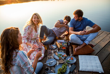 Group of friends having fun on picnic near a lake, sitting on pier eating and drinking wine.