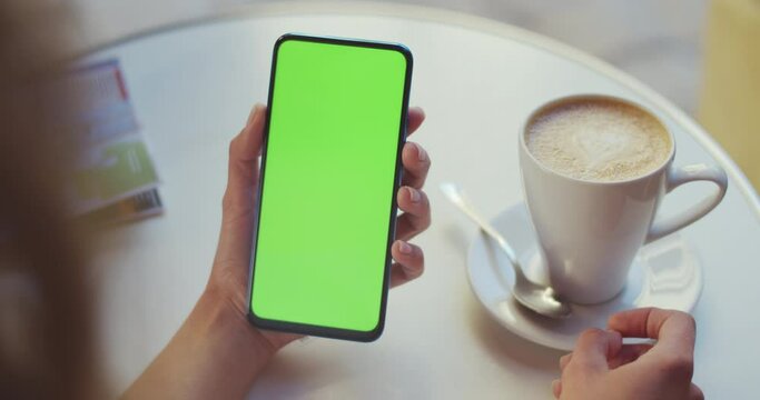 Crop view of female person traveler holding green screen smartphone while sitting at table with coffee cup and map on it. Concept of mockup and chroma key.