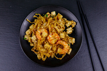 Stir-fried rice noodles Pad Thai with prawns and tofu in a plate on black slate background. Top view. Thai food