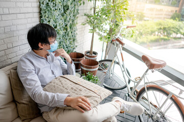 Asian man in casual clothing wearing protective face mask sitting on comfortable sofa with his bicycle. Quarantine at home concept