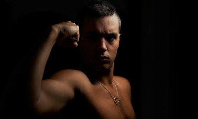 Obraz na płótnie Canvas Young muscular guy looking straight ahead and showing his arm. Shirtless muscular teenage boy on black background. Young shirtless brunette on black background. Athlete and short hair with pendant