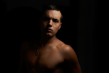 Fototapeta premium muscular young guy looking straight ahead. Shirtless muscular teenage boy on black background. Young shirtless brunette on black background. Athlete without a shirt and short hair with pendant.