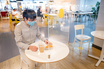 Asian man wearing face mask sitting in table shield partition preparing for eating croissant and coffee at cafe restaurant. Preventing COVID-19 (Coronavirus) infection. Social distancing concept