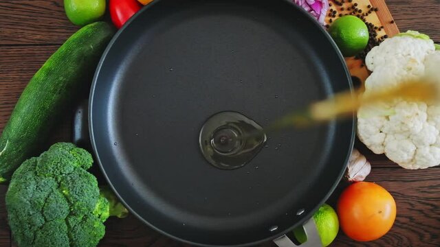 Slow motion  of  oil pouring into a frying pan. Cook pours sunflower oil into a frying pan, top view. Kitchen pan stands on brown boards, vegetables around it, and oil is poured into it for frying.  