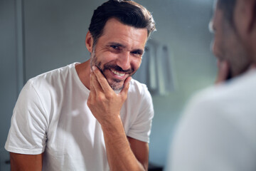 An young smiling man is applying after cleaning his face day or night cream to take care of skin behind a mirror in a bathroom.