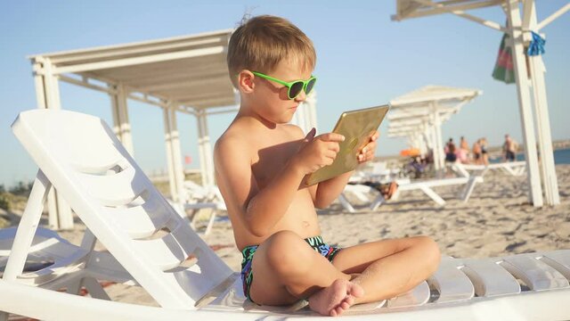 child is sitting on a sun lounger on the beach with a tablet in his hands. A boy sitting on the beach takes a photo on a tablet.