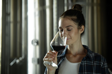 Authentic shot of happy successful female winemaker is tasting a flavor and checking red wine quality poured in transparent glass  in a wine cellar.
