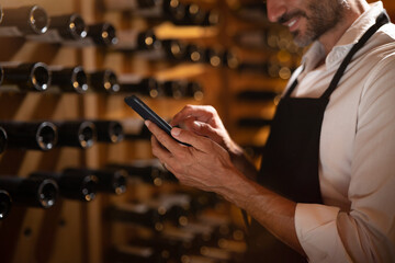 Modern farmer or winemaker is using winery online commerce applications on a smartphone for checking customer service and selling orders summary of his wine production in a wine cellar.