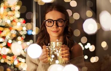 christmas, holidays and people concept - young woman in glasses with garland lights in mason jar...