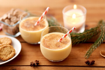 christmas and seasonal drinks concept - glasses of eggnog with oatmeal cookies, candy canes, sugar, fir tree branches and candle burning on wooden background