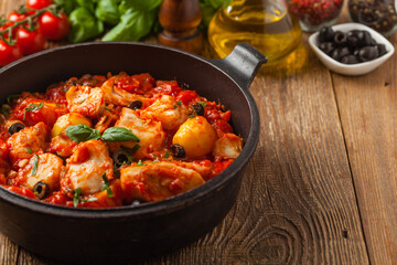 Traditionally prepared cod in Spanish. Served in tomato sauce with boiled potatoes, olivki and basil. Front view.