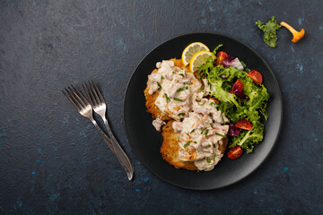 Fried cod, served on potato pancakes with mushrooms sauce and salads. Portion on a black plate. Dark background.