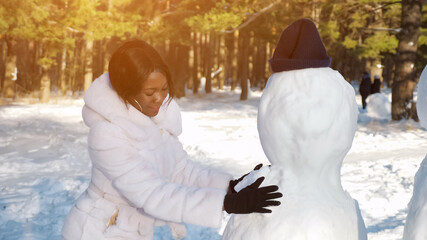 African-American woman in fur coat and black gloves makes snowman among winter nature in sunny...