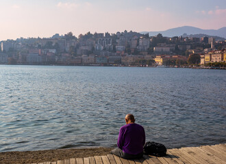 Fototapeta premium man relaxes sitting on wooden pier with panoramic view of Lugano lake, Lugano city and surrounding mountains. Sunset landscape.Lugano, Ticino canton, Switzerland.