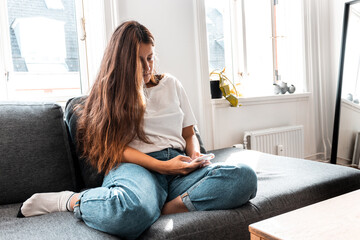 Mujer joven sentada en un sofa enviando mensajes de texto en tiempos de corona virus - quedarse en...