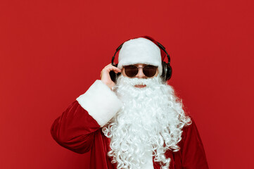 Studio closeup portrait of cool Santa Claus in sunglasses listening to music in headphones and looking at camera on red background. The cool modern Santa loves to listen to music. Xmas. Copyspace