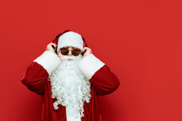 Fototapeta na wymiar Cool Santa Claus in sunglasses listens to music in headphones and has fun isolated on red background. Christmas and New Year concept. Portraits of modern Santa. Xmas