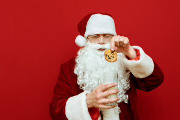 Beautiful Santa eating cookies and milk on a red background, puts cookies in glass. Christmas and New Year concept. Santa Claus and cookies with milk. Isolated. Background