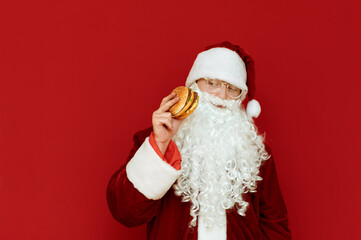 Fototapeta na wymiar Portrait of serious Santa Claus in glasses and white beard isolated on red background with burger in hand, looking aside for fast food with serious face. Funny Santa Claus eats fast food. Christmas