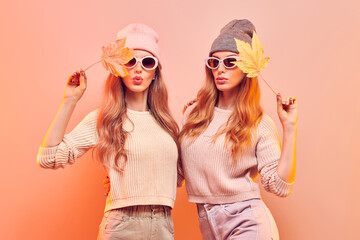 Autumn. Two fashionable hipster woman sisters in Trendy fall outfit, stylish hair, makeup. Friends in jumper, fashion jeans having fun. Beautiful girl in autumnal beanie hat with maple leaf