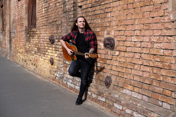 Fototapeta na wymiar Young male with long hair and tattoo play on acoustic guitar outdoors on the street 