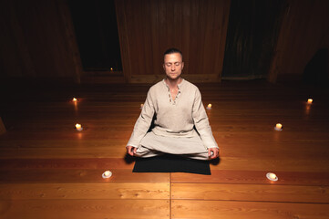 A portrait of an attractive Caucasian man sits in cotton robes in a lotus pose in a dark wooden...