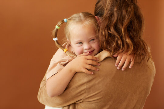 Back view portrait of mother holding happy daughter with down syndrome while standing against plain brown background in studio