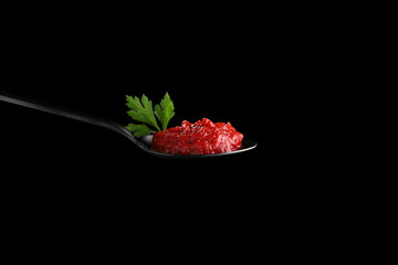 Red tomato paste on a black spoon, garnished with parsley leaves. On a black background. An ingredient for many dishes.