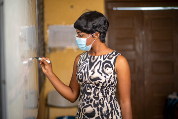 portrait of beautiful African teacher in face mask, writing on marker board after lock down, black tutor in short hair in lecture room-education concept 