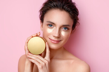 Woman with eye patches showing jar with cosmetic cream. Photo of beautiful woman on pink background. Beauty and skin care concept