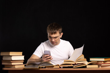 YOUNG MAN IS SITTING AT THE TABLE WITH BOOKS AND LOOKING INTO THE SMARTPHONE. DISTANCE LEARNING in quarantine. accountant works in an office against a black wall.