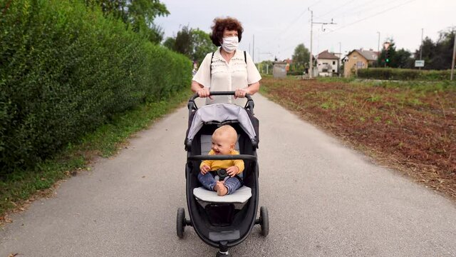 A grandmother with a medical mask pushing an upset baby boy in a stroller on a street. Covid-19 separating families.