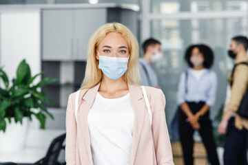 Modern manager returns to work after quarantine. Focus on attractive blonde woman in protective mask and business suit, employees on blurred background