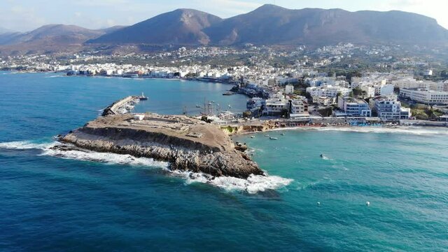 Scenic aerial view of small tourist town and half-island, crystal waters of Aegean sea. Hersonissos little port shelter at North Shore of Crete Island. End of summer season