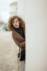 Obraz premium A portrait of a curly-haired young woman peeking out from behind a white pillar. The girl looks in surprise, putting her lips in a bow