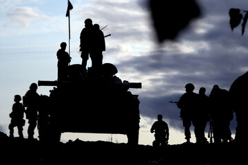 Armoured personnel carrier and a group of soldiers with defocused tree leaves in the foreground.