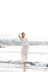 Fototapeta na wymiar Happy young pregnant woman in a long dress walks on the sea beach.
