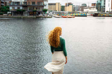 beautiful red haired girl standing on a pier facing water, back to camera, cityscape in background